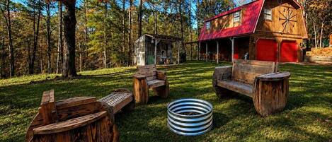 Un chalet rustique avec un toit rouge, entouré d’une cour verdoyante aménagée avec des meubles en bois et un foyer en métal.