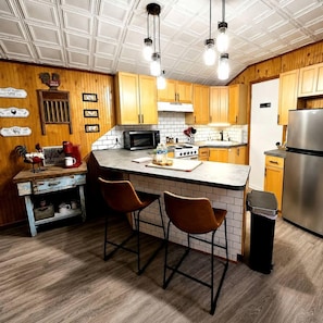 A kitchen with wooden cabinets, a central island with bar stools, a stainless steel refrigerator, and a microwave.