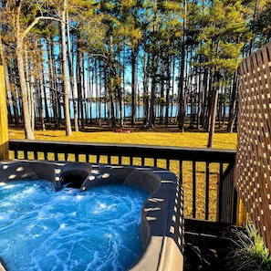 A hot tub surrounded by a wooden deck, with a view of a forest and a body of water in the background.