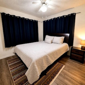 A bedroom with a bed, dark curtains, a ceiling fan, a nightstand, and a patterned rug.
