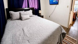 A neatly made bed with a headboard, a ceiling fan, a flat-screen TV mounted on the wall, and a patterned rug on the floor.