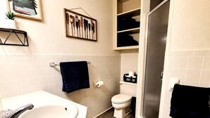 A bathroom with a white sink, toilet, and shower area. There is a towel rack with a dark towel, a framed picture of paddles, and a small plant on the sink.