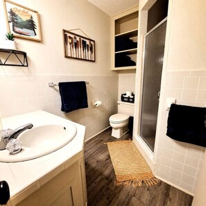A bathroom with a white sink, toilet, and shower area. There is a towel rack with a dark towel, a framed picture of paddles, and a small plant on the sink.