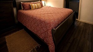 Een slaapkamer met een bed, een kaptafel, een nachtkastje met een lamp, een schilderij aan de muur, en een deur naar een andere kamer.