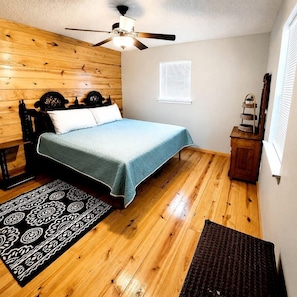 Un dormitorio con cama, paredes de madera, ventilador de techo, ventana con persianas y un pequeño armario de madera.