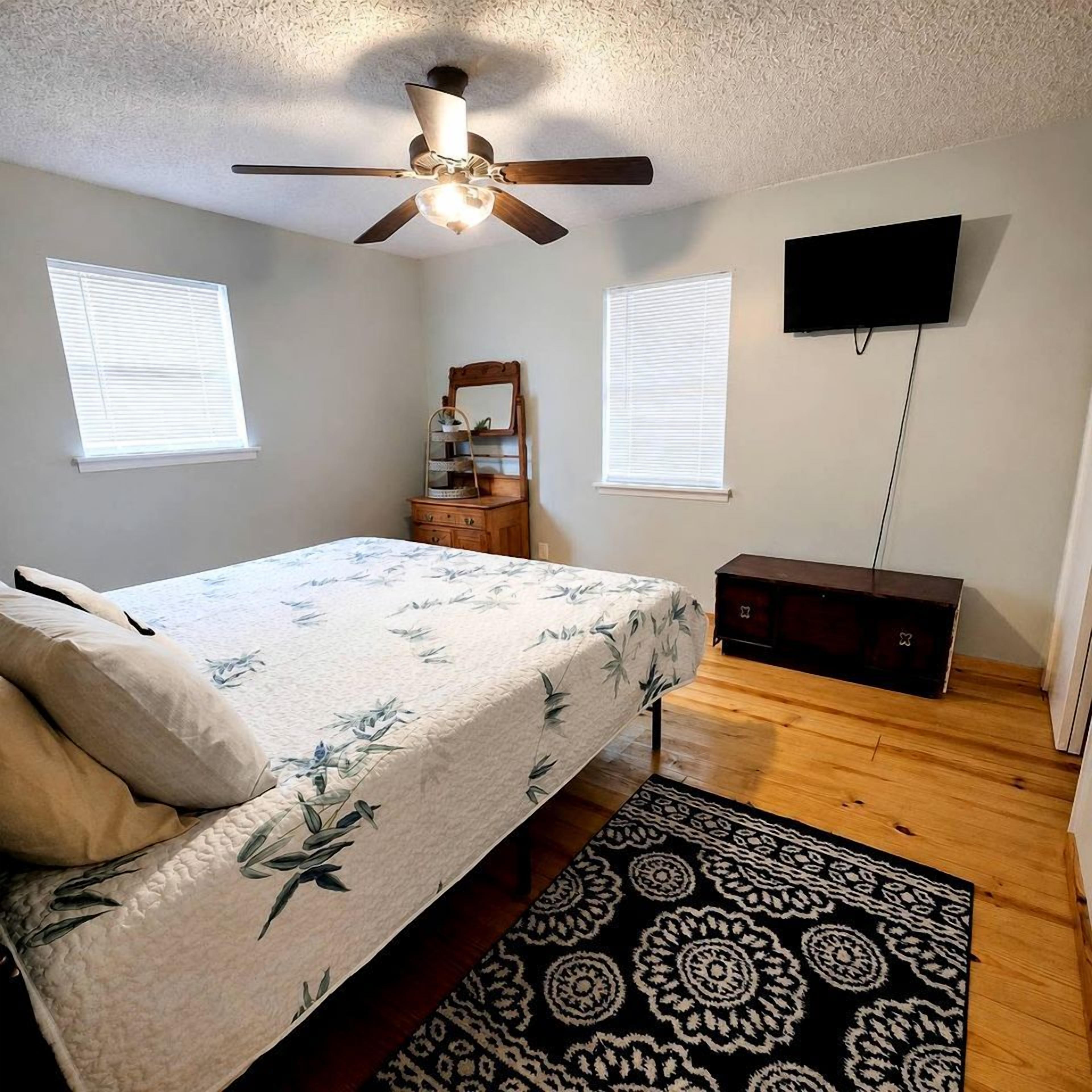 Quarto com cama, ventilador de teto, TV na parede e cômoda.