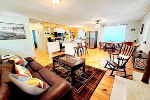A living room with a brown sofa, a coffee table, and a rocking chair. There is a kitchen area with a refrigerator and a dining table with chairs.