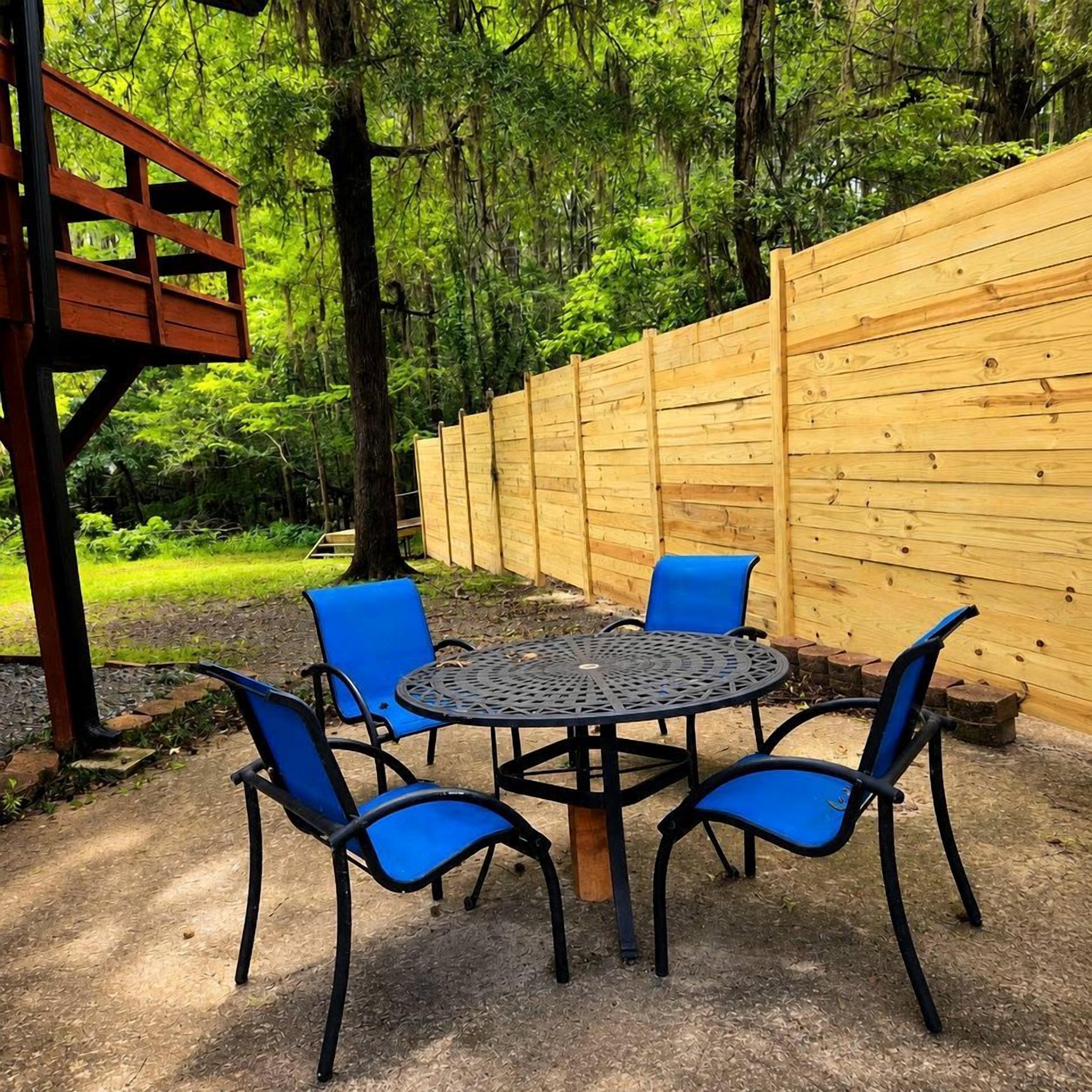An outdoor seating area with a round metal table and four blue chairs, surrounded by a wooden fence and trees.