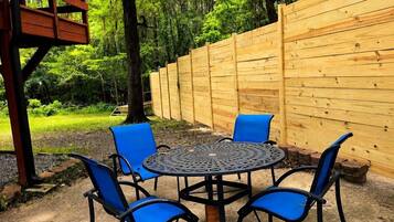 An outdoor seating area with a round metal table and four blue chairs, surrounded by a wooden fence and trees.
