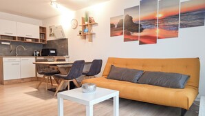 Een moderne woonkamer met een bank, twee fauteuils, een klein tafeltje en een keukenhoek met kastjes en een spoelbak.