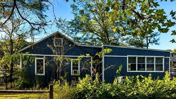 A blue building with a sloped roof, surrounded by greenery and a wooden fence.