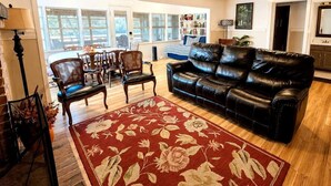 A living room with a leather sofa, wooden chairs, a red floral rug, and a ceiling fan.