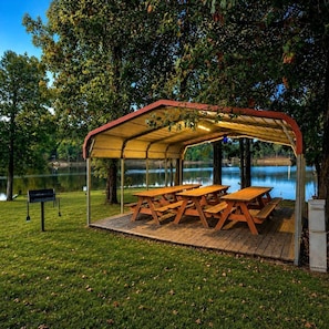 A covered picnic area with tables and benches, surrounded by trees and a body of water.