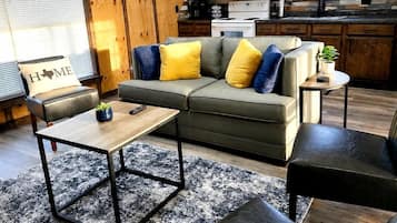 Ein gemütliches Wohnzimmer mit Sofa, Couchtisch und Hocker, eine Küche mit Holzschränken und Geräten sowie ein Fenster mit Jalousien.