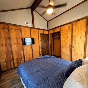 A bedroom with a wooden paneled wall, a ceiling fan, a flat-screen TV, and a bed with blue bedding.
