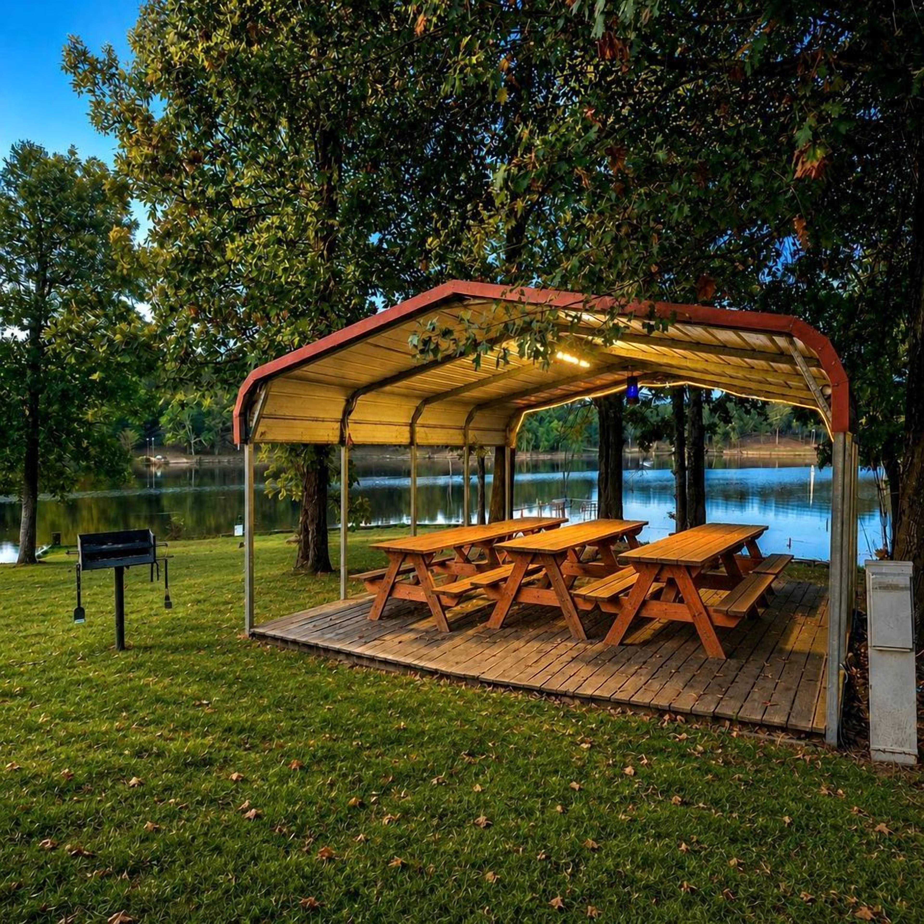 Un espace pique-nique abrité, équipé de tables et de bancs, entouré d’arbres et bordé par un plan d’eau.
