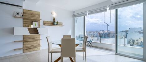 Un comedor moderno con una mesa y sillas de vidrio, estantes de madera y vistas a la ciudad a través de grandes ventanales.