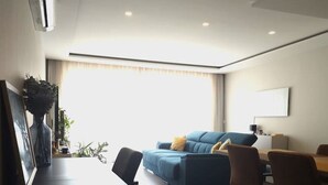 A modern living room with a sofa, dining table, and chairs. There is a window with curtains, a painting on the wall, and a plant on the side table.