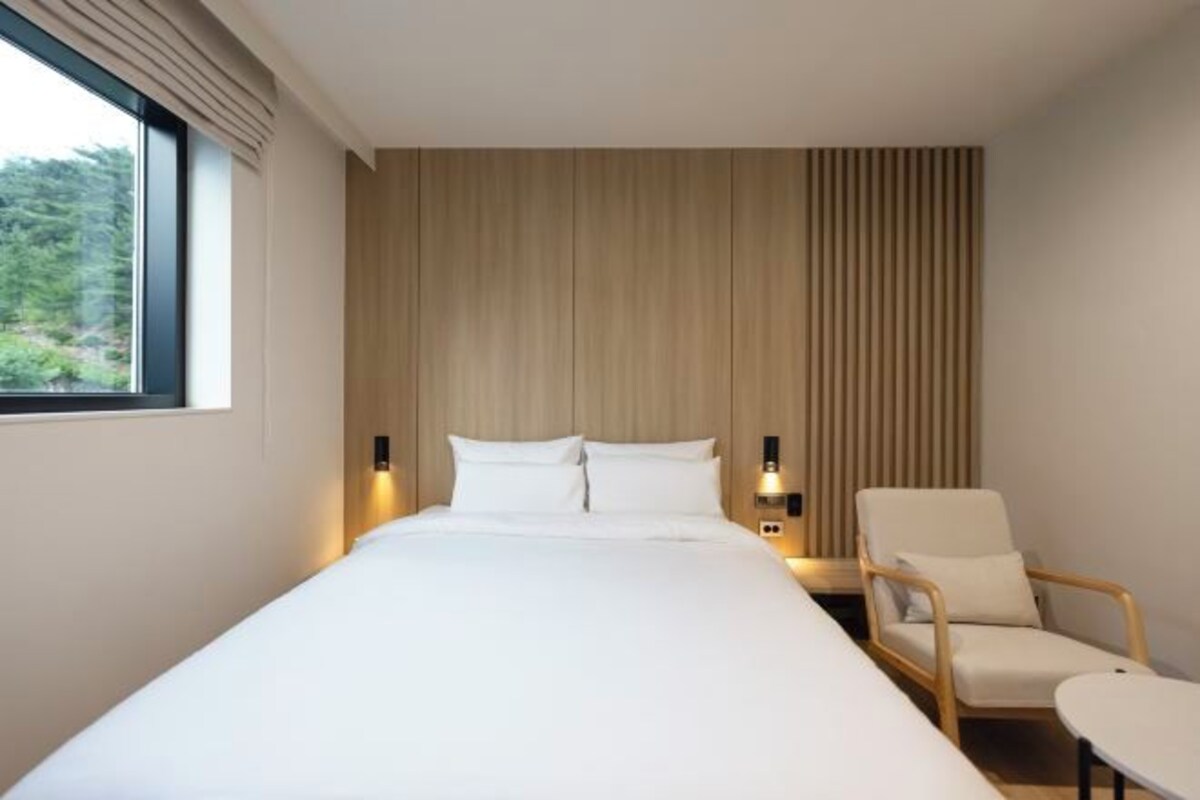 A modern hotel room with a large bed, a chair, a small table, and a window with a view of greenery.