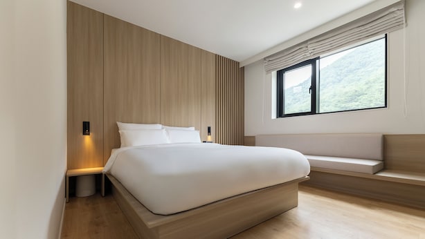 Un dormitorio moderno con una cama grande, una mesita de noche, una lámpara de pared y una ventana con vistas a la vegetación.