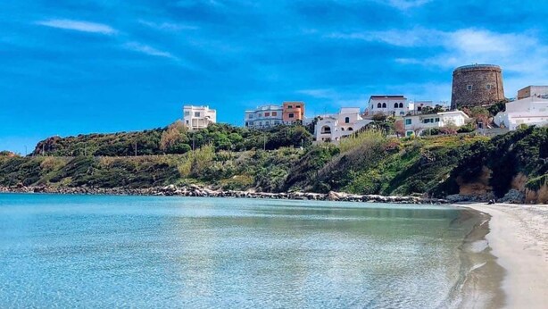 A coastal town with buildings on a hillside overlooking a clear beach. - Casa Susanna – Your Retreat in Calasetta: Sea, Culture, and Relaxation (Calasetta)
