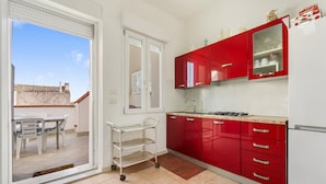 A kitchen with red cabinets, a white refrigerator, and a sliding glass door leading to a patio area with outdoor furniture. - Casa Susanna – Your Retreat in Calasetta: Sea, Culture, and Relaxation (Calasetta)