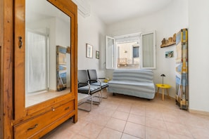 A room with a wooden wardrobe, a bed with striped bedding, a chair, a small table, and a window with a view of the outside.
