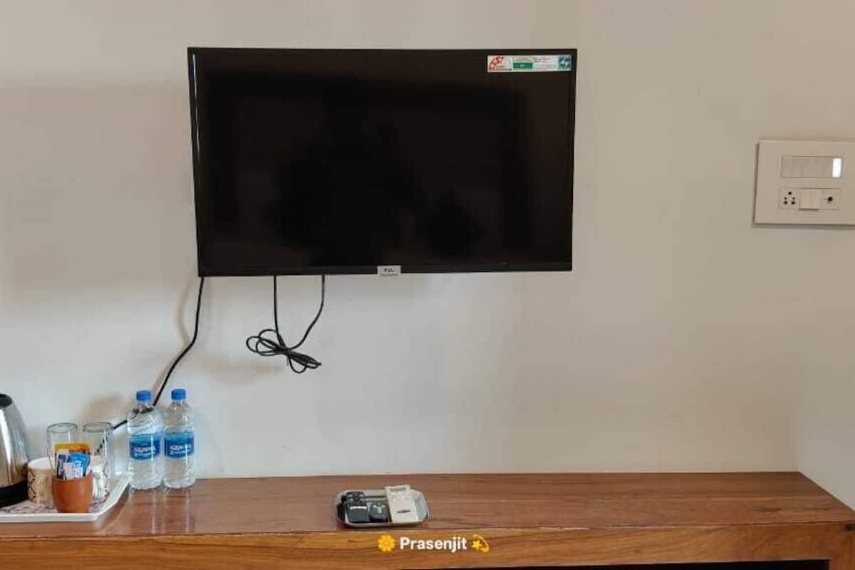 Habitación de hotel con televisor de pantalla plana en la pared, un enchufe, una mesa de madera con una tetera, vasos y botellas de agua.