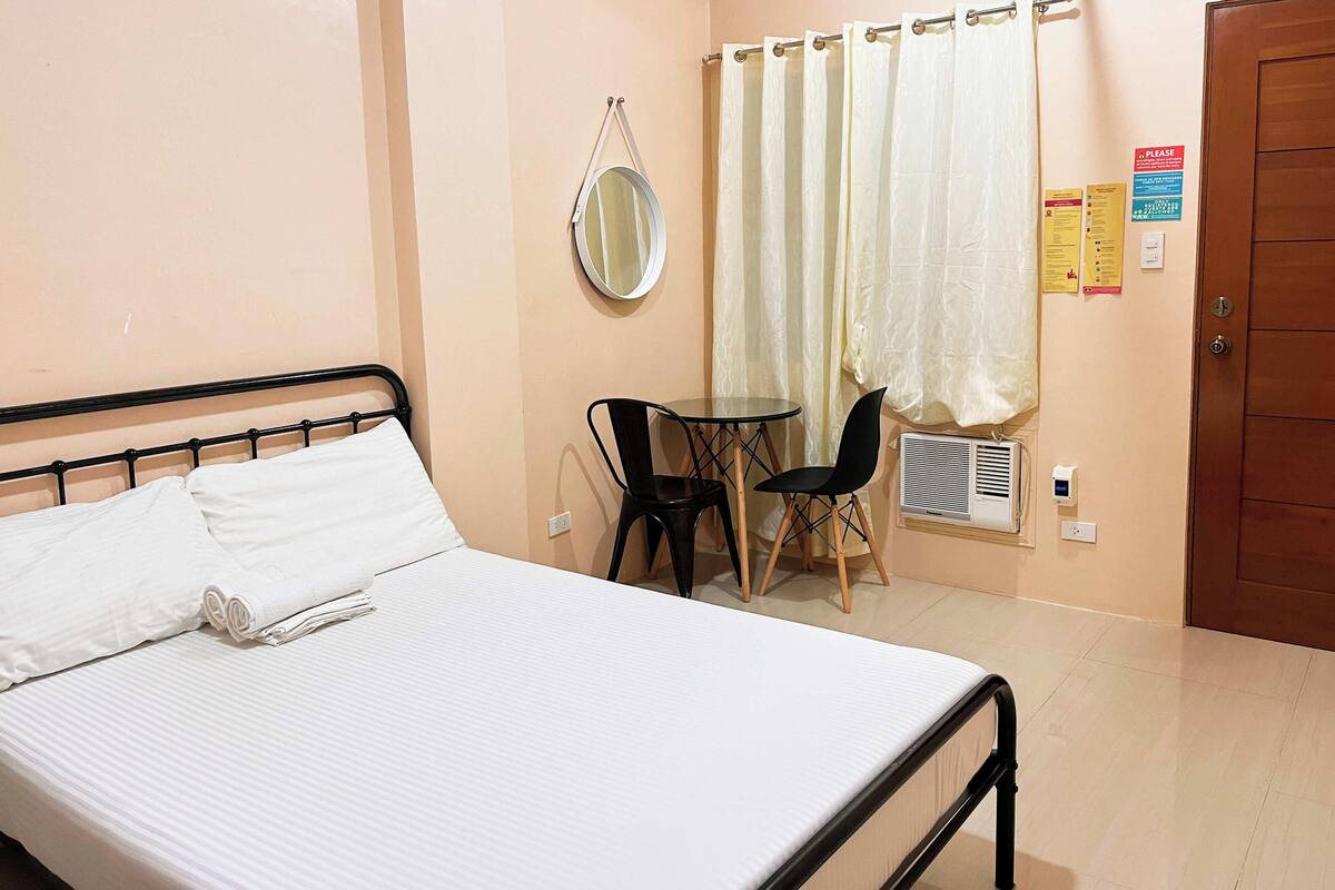 A neatly made bed with white linens, a small round table with two chairs, a mirror on the wall, and a window with a curtain.