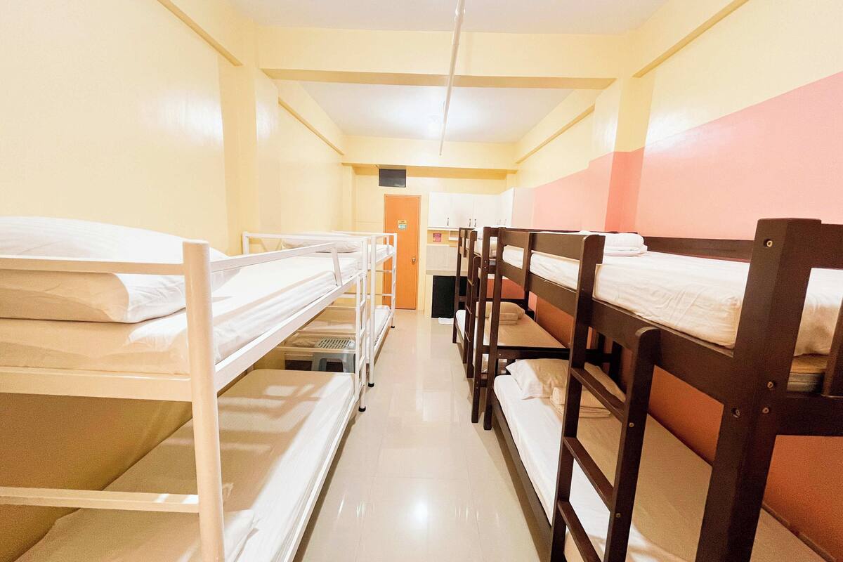 A dormitory room with bunk beds arranged in two rows, a kitchenette area, and a door leading to another room.