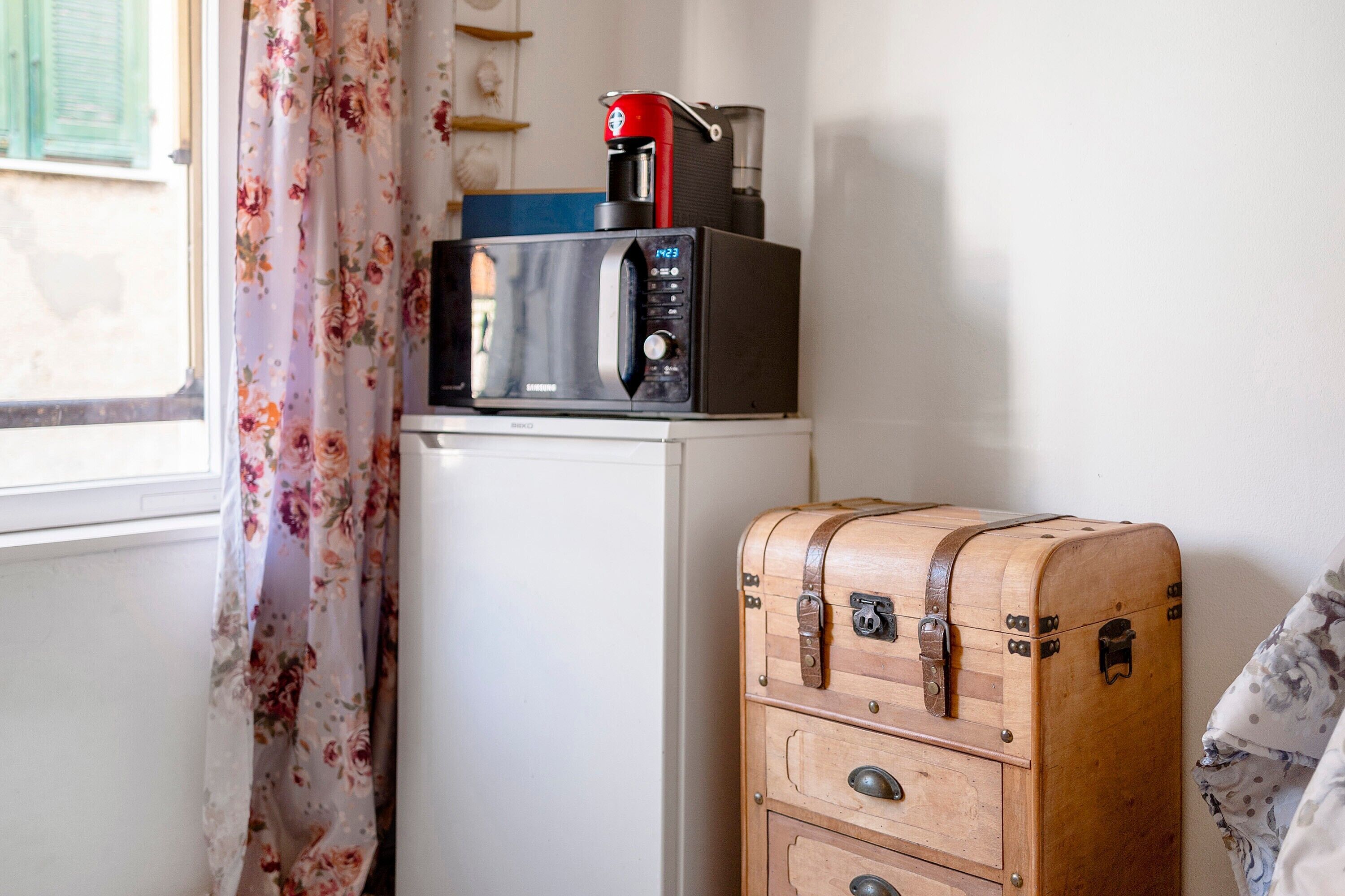 A microwave on a white appliance, a wooden suitcase, and a window with floral curtains.