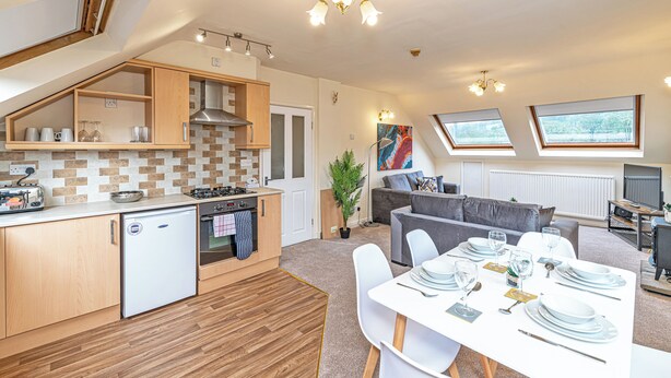 A compact kitchen with a refrigerator, stove, and wooden cabinets. There is a dining area with a table set for four, and a living area with a sofa and a television. - Scosthrop - Bridge End Mill By Guestz (Settle)