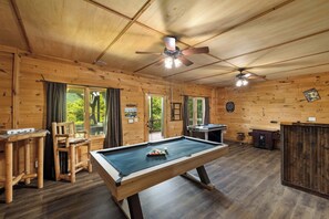 Ein Holzinnenraum mit Billardtisch, Deckenventilatoren und Blick auf Grünfläche im Freien.