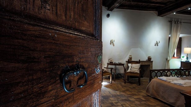 Una habitación rústica con puerta de madera, una cama, una silla, una mesita y una lámpara.