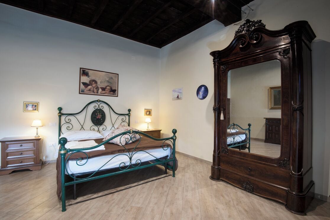 A bedroom with a metal bed frame, a wooden nightstand, a bedside table with a lamp, a large mirror, and a wall-mounted picture.