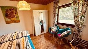 A cozy room with a bed, two chairs, a wooden bench, a window with curtains, and a door leading to another room. - Cozy bungalow at the foot of the Wasserkuppe for up to 4 people (Dipperz)