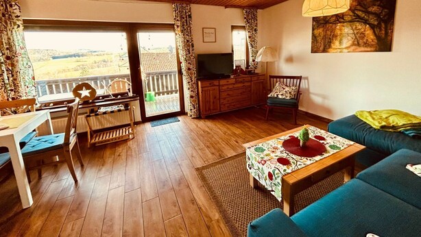 A cozy living room with a wooden ceiling, a sofa, a table with a floral tablecloth, and a television on a wooden cabinet. - Cozy bungalow at the foot of the Wasserkuppe for up to 4 people (Dipperz)