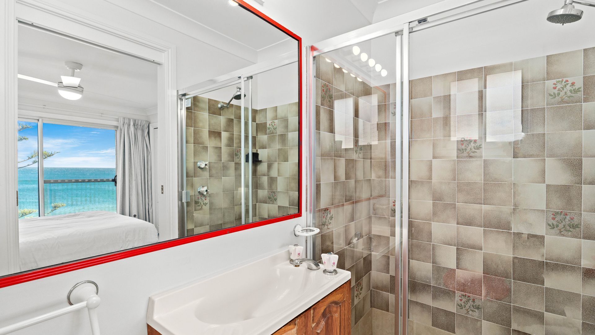 A modern bathroom with a large mirror, a shower with glass doors, and a view of the ocean through the window.