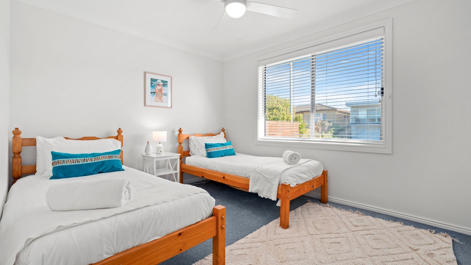 A bedroom with two wooden beds, white bedding, a small bedside table with a lamp, a framed picture on the wall, and a large window with blinds.
