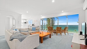 A spacious living area with a large window overlooking the sea, a kitchen, and a seating area with wicker chairs. - Forresters Dunes - Upstairs (Forresters Beach)