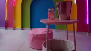 Une pièce avec une arche néon aux couleurs vives, une table ronde surmontée d’un seau en métal, un pouf rose et un panier blanc.