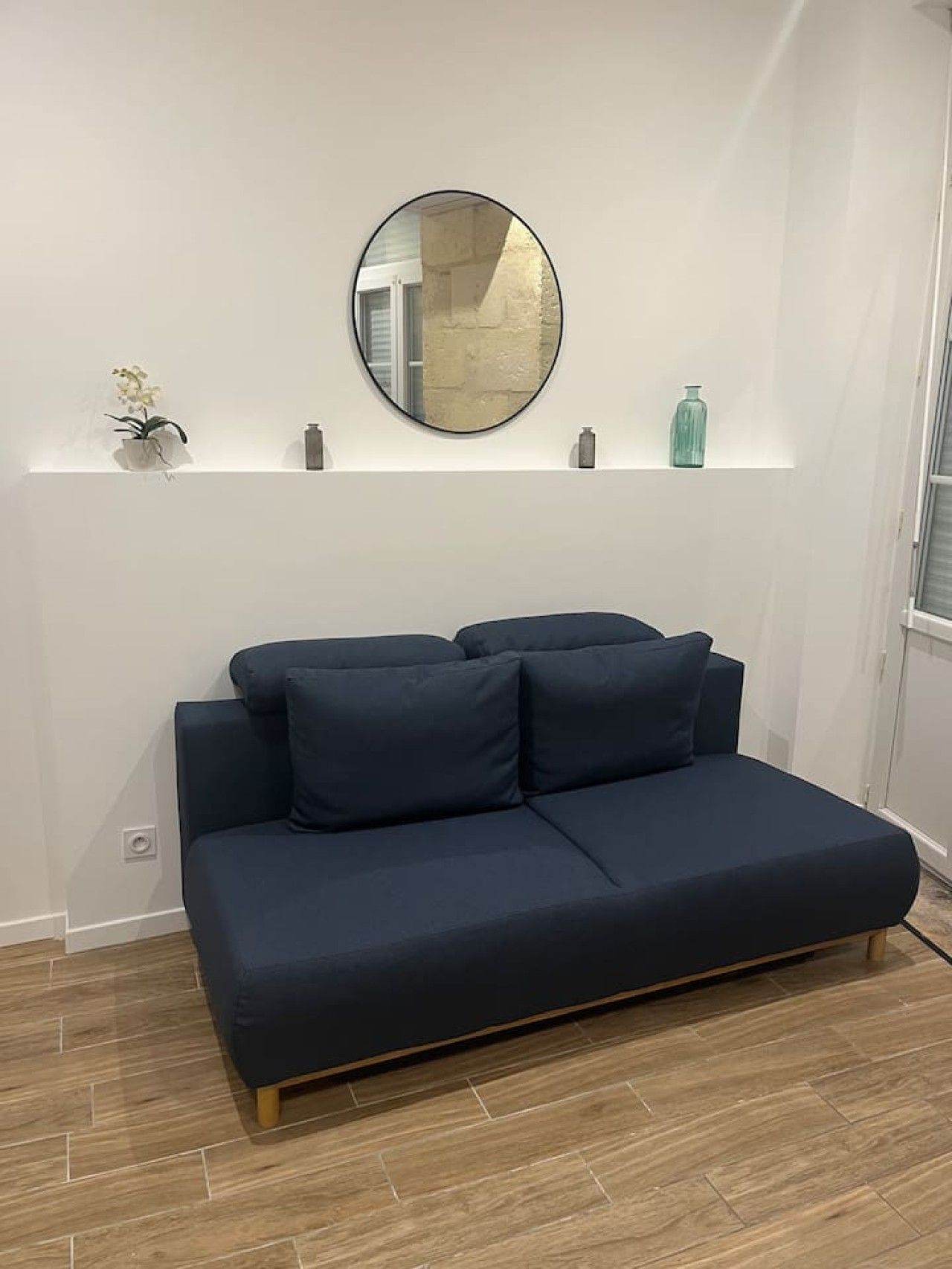 Een moderne woonkamer met een donkerblauwe bank, een ronde spiegel aan de muur, en een klein plankje met decoratieve objecten.