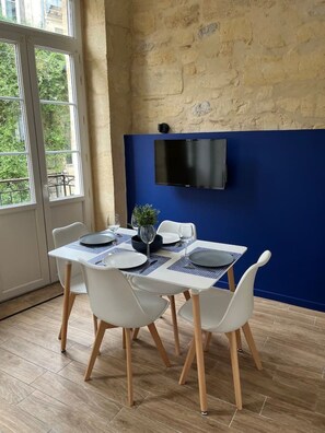 Een eetgedeelte met een tafel gedekt voor twee personen, witte stoelen, een blauwe wand en een wandgemonteerde televisie.