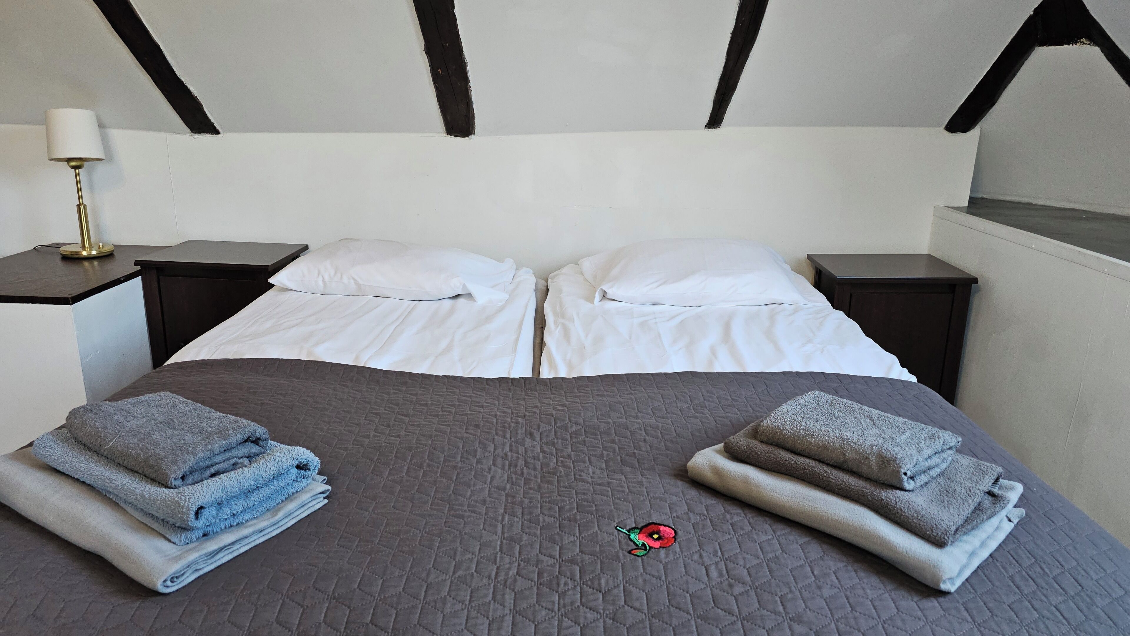 Una cama bien tendida con sábanas blancas y colcha gris, flanqueada por dos mesitas de noche con lámparas, en una habitación de diseño moderno y sencillo.