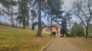 A paved pathway leading to a building with a red roof, surrounded by trees and grass. - Villa Podkarpacka (Kudowa Zdroj)