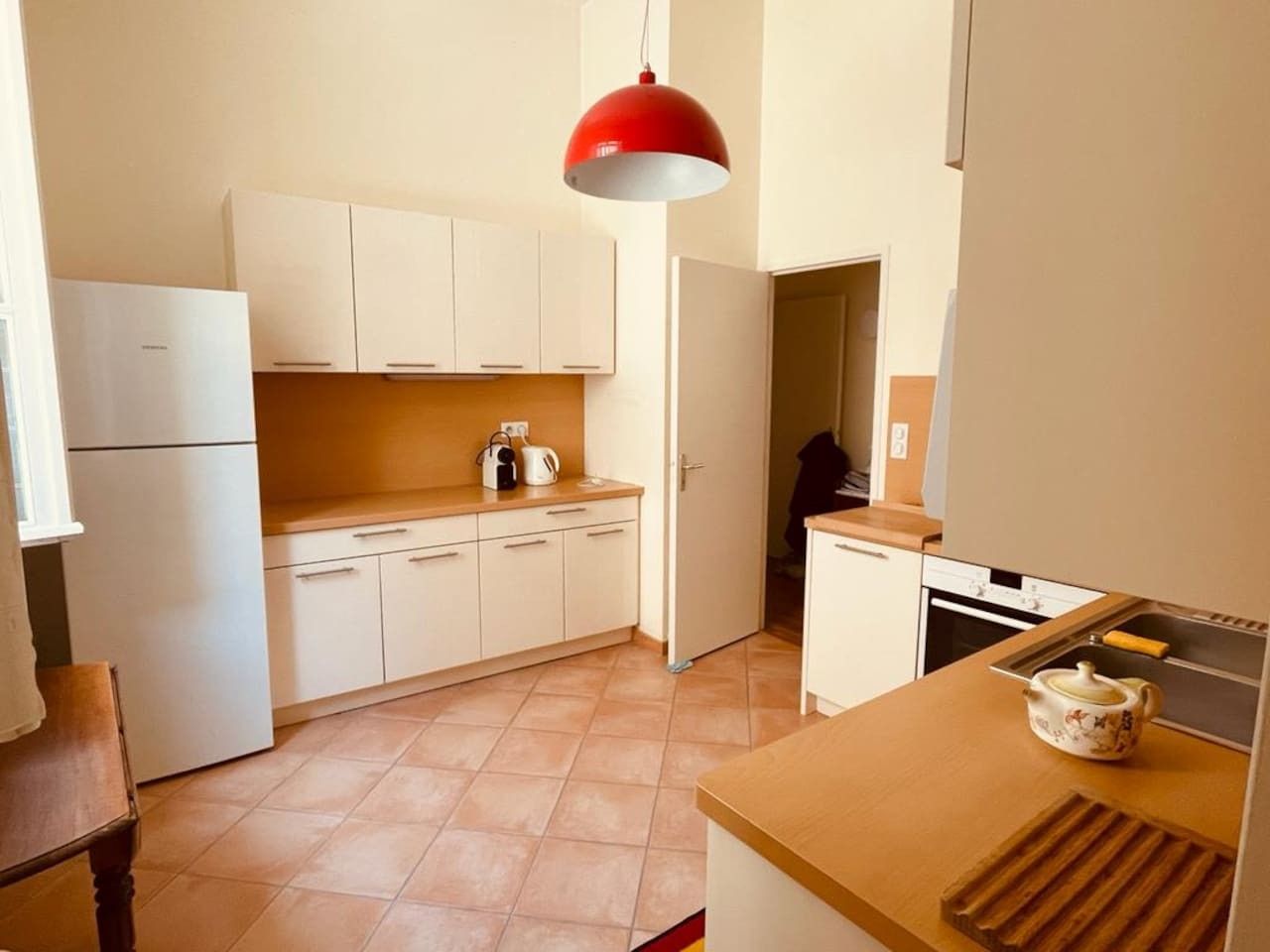 Een kleine keuken met witte kasten, een rode hanglamp en een klein aanrecht met een theepot.