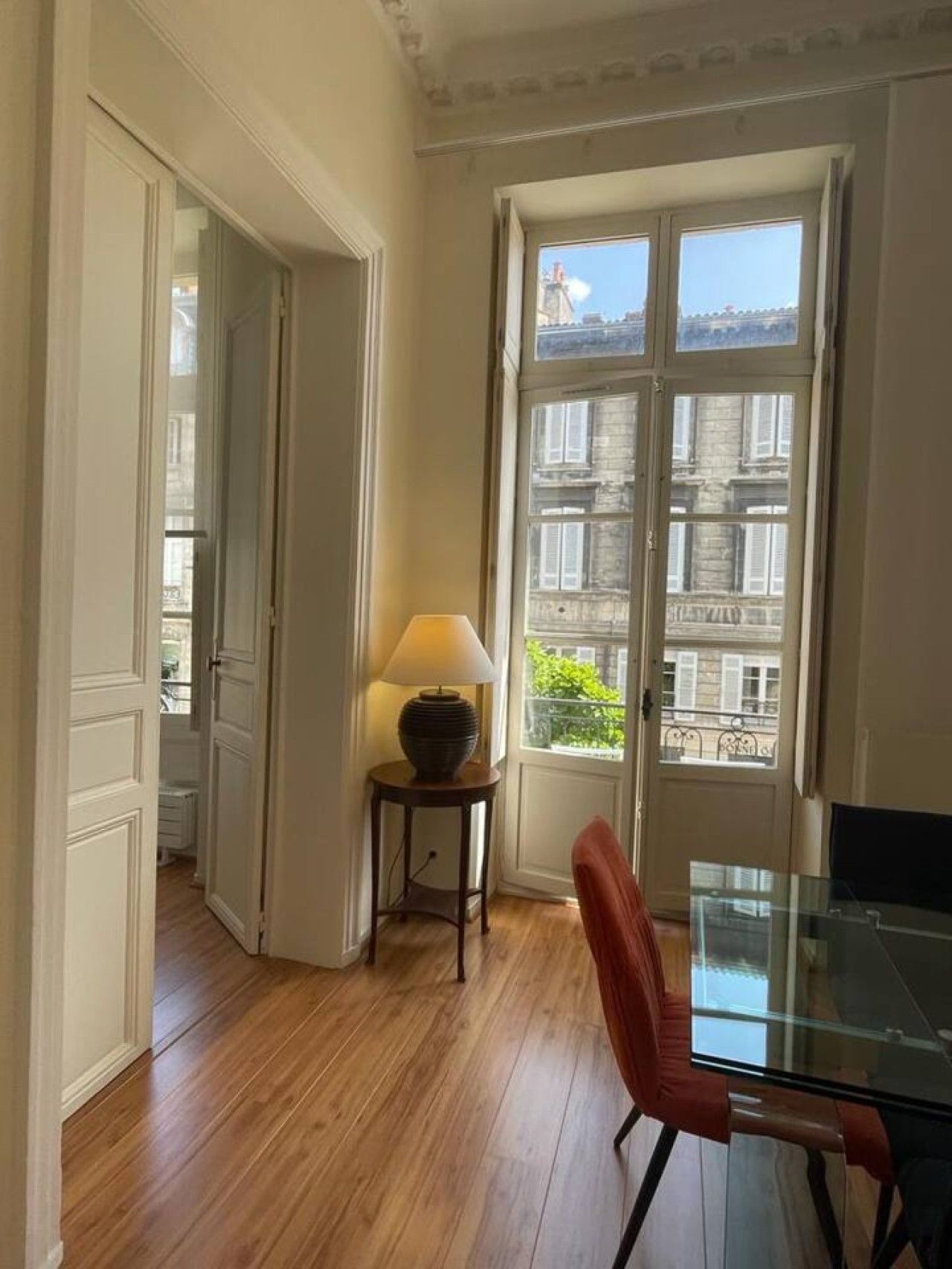 Een kamer met een houten vloer, een glazen tafel, een rode stoel, een lamp op een bijzettafeltje en een deur die leidt naar een buitenbalkon met uitzicht op gebouwen.