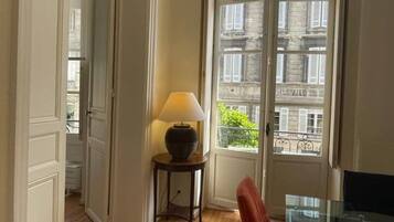 Een kamer met een houten vloer, een glazen tafel, een rode stoel, een lamp op een bijzettafeltje en een deur die leidt naar een buitenbalkon met uitzicht op gebouwen.