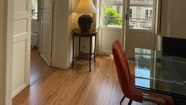 A room with a wooden floor, a glass table, a red chair, a lamp on a small table, and a large window with a view of buildings. - Elegance et confort au centre de Bordeaux (Bordeaux)