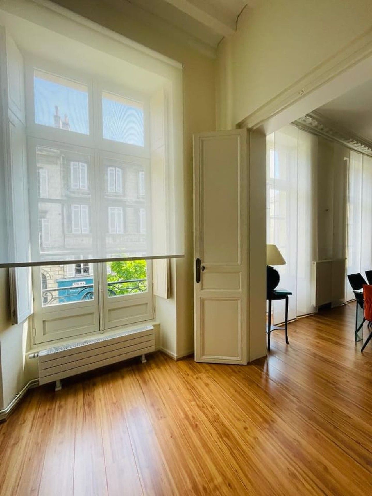 Een kamer met een groot raam, een radiator, een deur en een stoel.